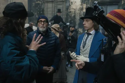 Grupa ludzi na planie filmowym, w tym reżyser w czapce i młody aktor w niebieskim fraku.