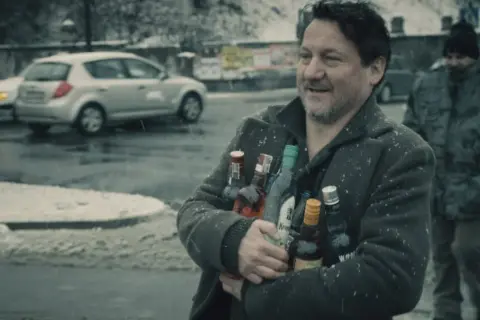Mężczyzna z uśmiechem niesie kilka butelek alkoholu pod mocnym aniołem obsada. W tle śnieg i samochody.