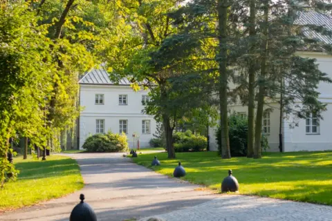 Biały dworek otoczony zielenią, z alejką i ozdobnymi kulami.