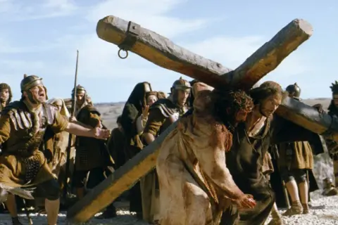 Jezus w koronie cierniowej niesie krzyż, otoczony przez rzymskich żołnierzy.