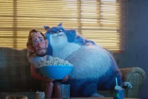 Wielki, niebieski kot przytula zmartwionego mężczyznę, który trzyma miskę popcornu. To scena z filmu "Jak pies z kotem", gdzie relacje między zwierzętami a ludźmi bywają skomplikowane.