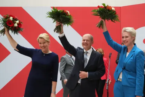 Uśmiechnięta Astrid Ann Marie Pollmann i politycy z bukietami kwiatów na tle czerwono-białej ściany.