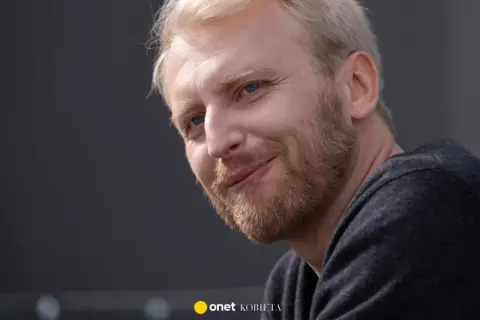 Uśmiechnięty Dobromir Dymecki z jasnymi włosami i niebieskimi oczami, w ciemnej bluzie.