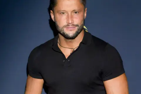 Piotr Mróz w czarnej koszulce polo, z zarostem i naszyjnikiem.