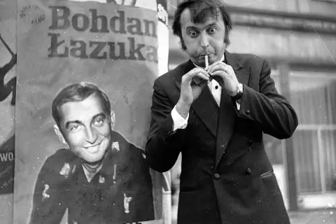 Bohdan Łazuka w garniturze, z papierosem w ustach, obok plakatu z jego podobizną.