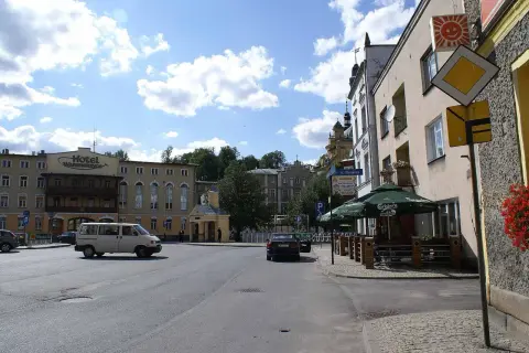 Plac miejski z hotelem, samochodami i kawiarnią pod zielonymi parasolami.