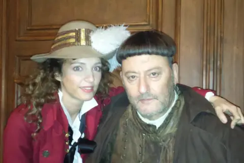 Goście, goście, obsada w historycznych strojach. Kobieta w kapeluszu z piórem i mężczyzna w brązowym płaszczu pozują do zdjęcia.