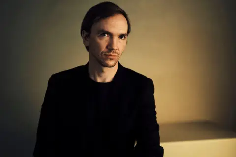 Portret Jana Komasy, reżysera w czarnym garniturze, na tle oświetlonej ściany.