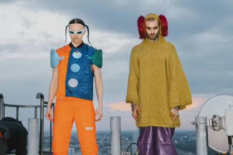 Tommy Cash i Quebonafide w ekstrawaganckich strojach na dachu. Jeden w pomarańczowo-niebieskiej kamizelce, drugi w żółtym swetrze i fioletowych spodniach.