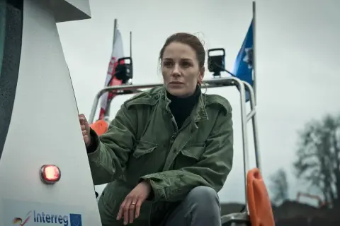 Katarzyna Wajda, aktorka, w zielonej kurtce, z polską flagą w tle, na tle pochmurnego nieba.