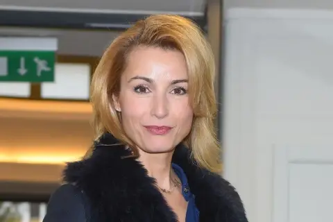 Joanna Brodzik uśmiecha się, ubrana w czarny płaszcz z futrzanym kołnierzem. W tle zielony znak ewakuacyjny.