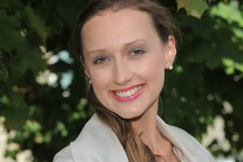 Uśmiechnięta Anna Gzyra w jasnej marynarce, z błyszczącymi kolczykami, na tle zielonych liści.