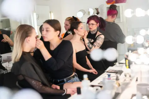 Charakteryzatorka filmowa pracuje nad makijażem modelki. W tle inne osoby przygotowują się do sesji.