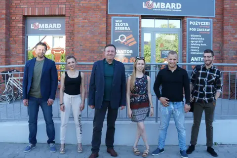 Aktorzy z lombardu pozują przed budynkiem.