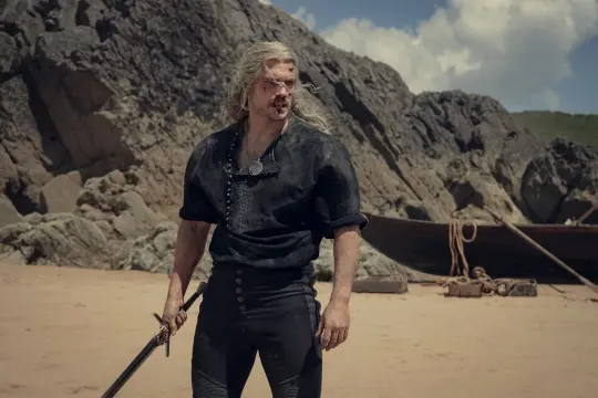 Wiedzmin Geralt z Rivii, ranny i zmęczony, stoi na piaszczystej plaży z mieczem w dłoni. W tle widać łódź i skaliste wybrzeże.