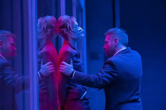 Mężczyzna w garniturze przytrzymuje kobietę z maską tlenową, która odbija się w lustrze. Scena z filmu "Tenet".