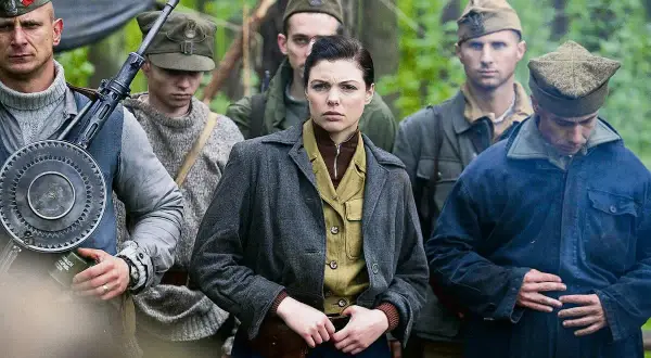 Grupa żołnierzy w lesie, w tym kobieta z pistoletem przy pasie. W tle widać broń maszynową. To scena z filmu "Czas honoru".