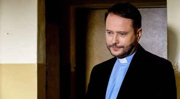 Aktor z obsady "Ksiądz Mateusz" w czarnym garniturze i błękitnej koszuli z białym kołnierzykiem, zamyślony.