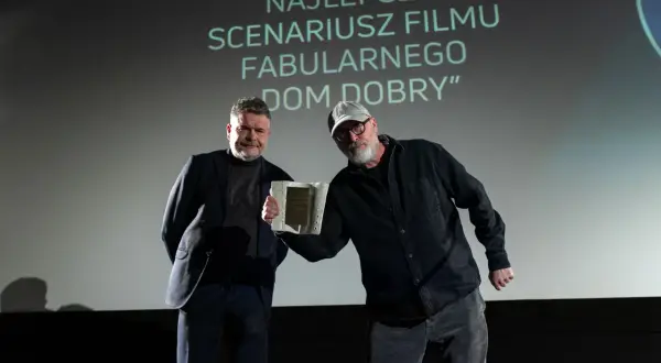 Dwóch mężczyzn na scenie, jeden odbiera nagrodę. Na ekranie napis "NAJLEPSZY SCENARIUSZ".