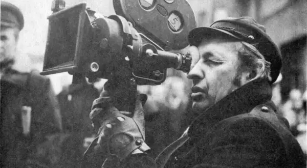 Operator filmowy w czapce i płaszczu patrzy przez wizjer kamery filmowej.