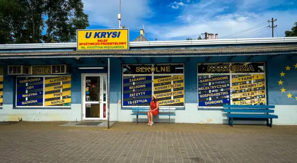 Dziewczyna w czerwonej sukience siedzi na ławce przed sklepem "U Krysi", gdzie można kupić wszystko, od zniczy po narzędzia.