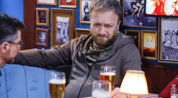 Tatusiowie serial obsada: Mężczyzna z brodą i w bluzie siedzi przy stole z piwem, w tle ściana z ramkami.