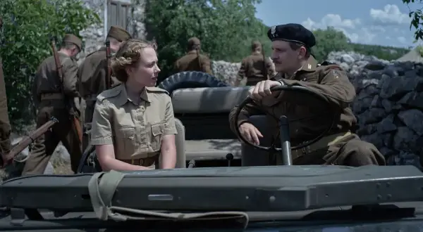 Żołnierze w mundurach z czasów II wojny światowej siedzą w jeepie na tle kamiennego muru.