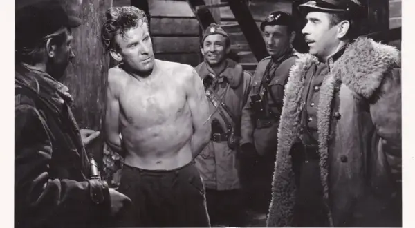 Czarno-białe zdjęcie przedstawia scenę z filmu: mężczyzna bez koszuli, z rękami związanymi, stoi przy drewnianym słupie, otoczony przez żołnierzy.