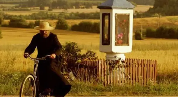 Mężczyzna w czarnym habicie i słomkowym kapeluszu jedzie rowerem obok kapliczki. Czuje się jak u pana boga w ogródku.
