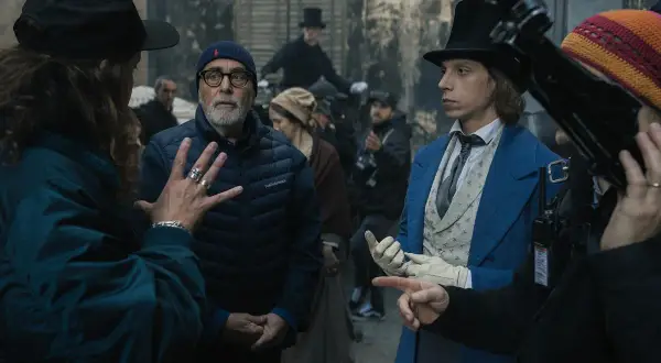 Grupa ludzi na planie filmowym, w tym reżyser w czapce i młody aktor w niebieskim fraku.