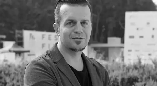 Czarno-białe zdjęcie Marcina Wrony, reżysera, w marynarce, na tle budynków i zieleni.