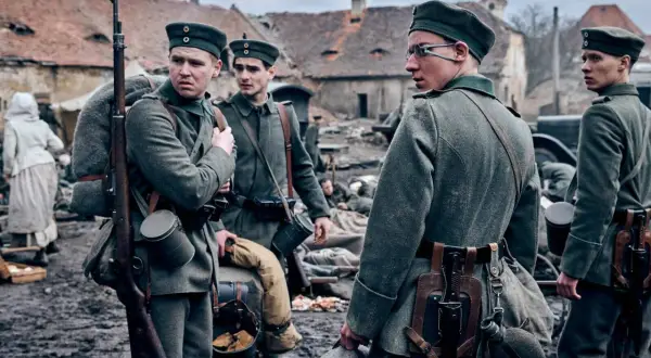 Żołnierze w mundurach z I wojny światowej, z karabinami i plecakami, stoją na błotnistym podwórzu. Na zachodzie bez zmian obsada.