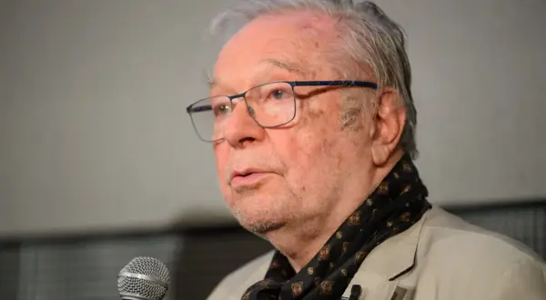 Portret starszego mężczyzny w okularach, mówiącego do mikrofonu, ubranego w jasną marynarkę i wzorzysty szalik.