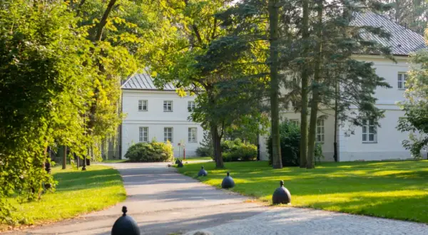 Biały dworek otoczony zielenią, z alejką i ozdobnymi kulami.