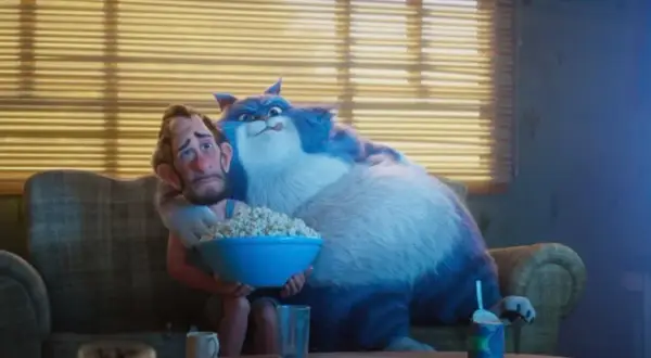 Wielki, niebieski kot przytula zmartwionego mężczyznę, który trzyma miskę popcornu. To scena z filmu "Jak pies z kotem", gdzie relacje między zwierzętami a ludźmi bywają skomplikowane.