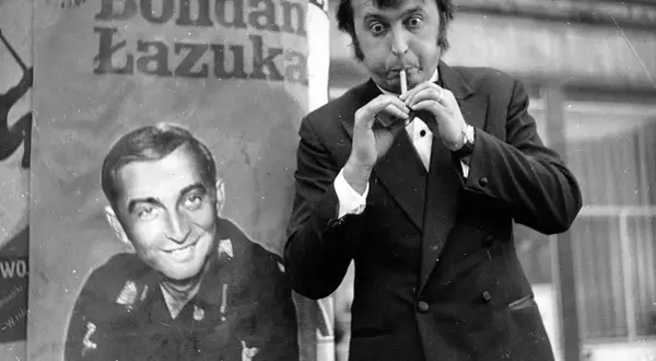 Bohdan Łazuka w garniturze, z papierosem w ustach, obok plakatu z jego podobizną.