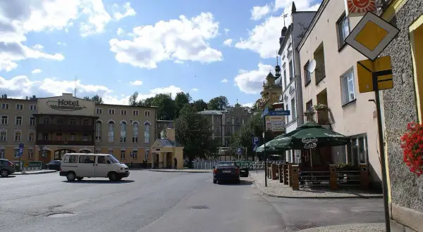 Plac miejski z hotelem, samochodami i kawiarnią pod zielonymi parasolami.