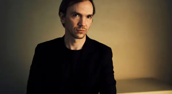Portret Jana Komasy, reżysera w czarnym garniturze, na tle oświetlonej ściany.