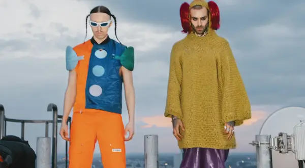 Tommy Cash i Quebonafide w ekstrawaganckich strojach na dachu. Jeden w pomarańczowo-niebieskiej kamizelce, drugi w żółtym swetrze i fioletowych spodniach.