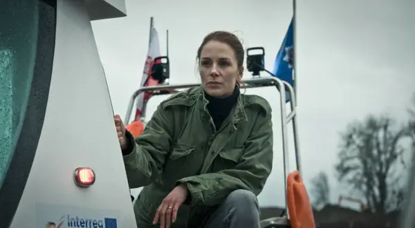 Katarzyna Wajda, aktorka, w zielonej kurtce, z polską flagą w tle, na tle pochmurnego nieba.