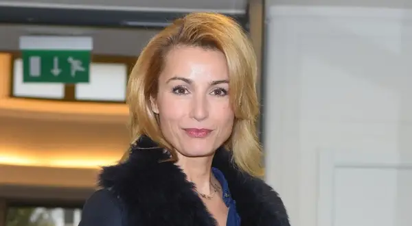 Joanna Brodzik uśmiecha się, ubrana w czarny płaszcz z futrzanym kołnierzem. W tle zielony znak ewakuacyjny.