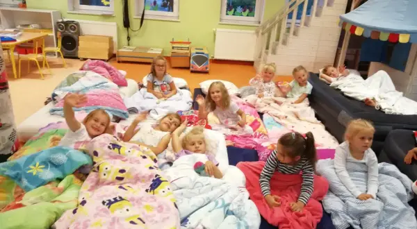 Dzieci leżą w śpiworach i kołdrach, gotowe na noc w przedszkolu. Niektóre machają do aparatu, inne patrzą z zaciekawieniem.