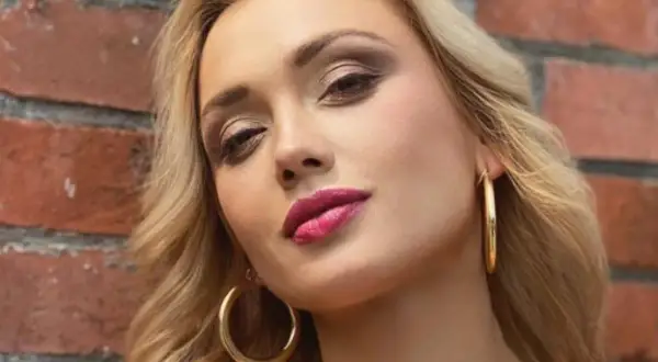 Dominika Skoczylas z błyszczącymi złotymi kolczykami i wyrazistym makijażem ust, na tle ceglanej ściany.