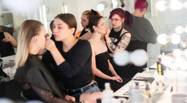 Charakteryzatorka filmowa pracuje nad makijażem modelki. W tle inne osoby przygotowują się do sesji.