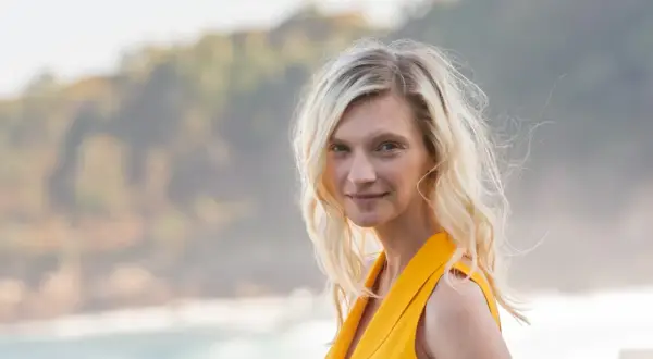 Agata Buzek w żółtej sukience, z falującymi blond włosami, uśmiecha się na tle morza i wzgórz.