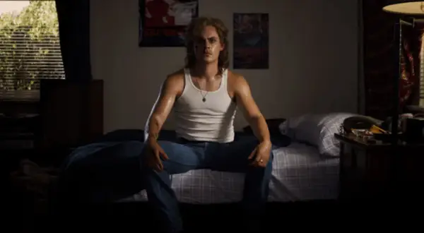 Billy z Stranger Things siedzi na łóżku w białej podkoszulce i dżinsach, w pokoju z plakatami na ścianie.