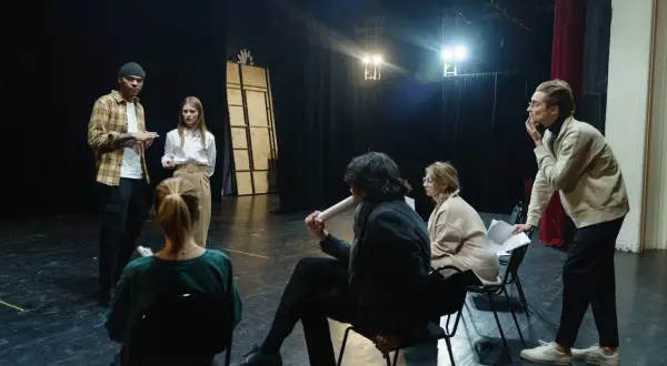 Grupa ludzi na scenie, dyskutująca o sztuce. Zastanawiają się, ile zarabia reżyser, analizując scenariusz.
