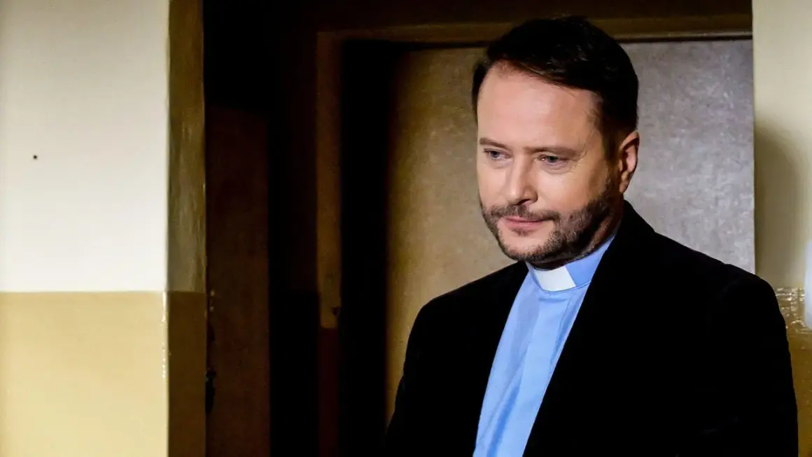 Aktor z obsady "Ksiądz Mateusz" w czarnym garniturze i błękitnej koszuli z białym kołnierzykiem, zamyślony.