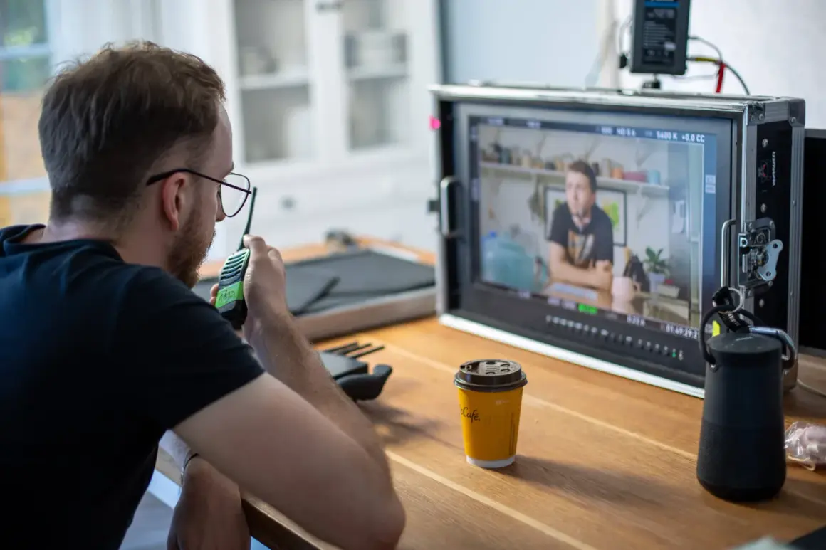 Producenci na planie filmowym czuwają nad realizacją, jeden z nich rozmawia przez krótkofalówkę, monitorując obraz na ekranie.