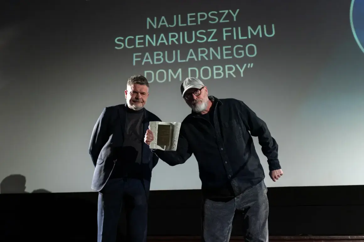 Dwóch mężczyzn na scenie, jeden odbiera nagrodę. Na ekranie napis "NAJLEPSZY SCENARIUSZ".